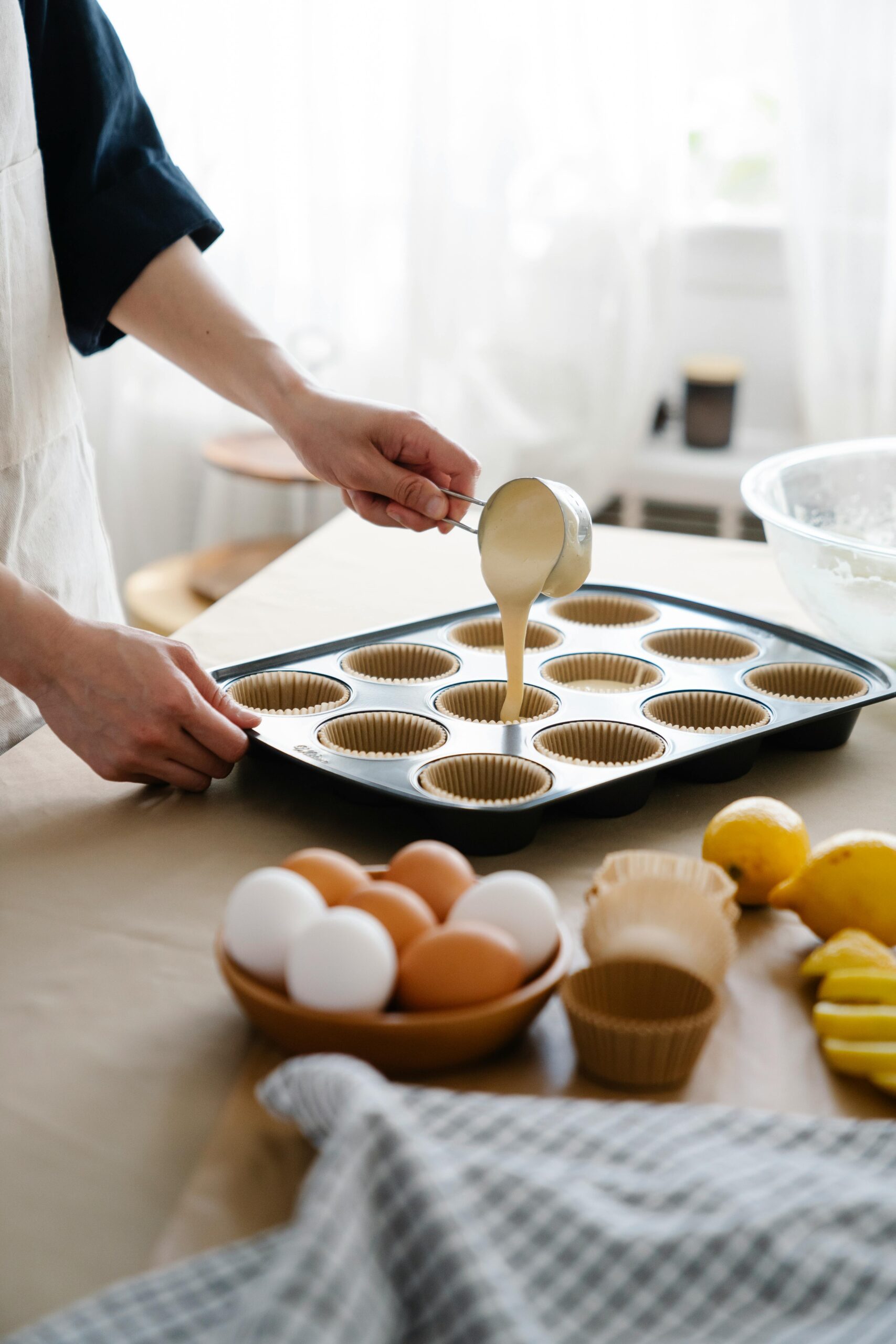 A baker pours cake batter into a muffin tray with eggs and lemons on a table.